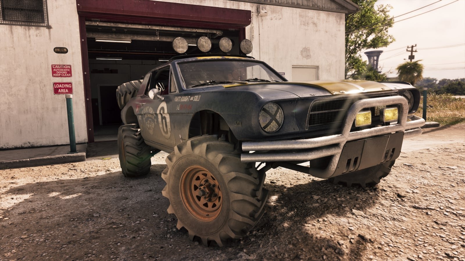 A battered blue and yellow off-road muscle car with giant tires and roof-mounted lights sits in front of a garage loading bay.